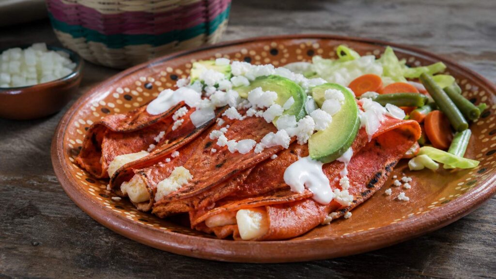 Enchiladas potosinas de masa rojiza con chile, queso, crema, aguacate y vegetales en plato de barro